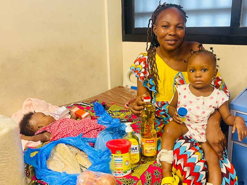 Eine afrikanische Frau umgeben von zwei kleinen Kindern und verschiedenen Lebensmitteln.