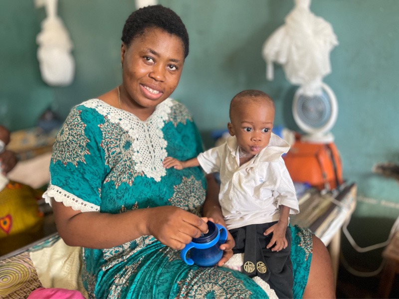 Eine Mutter hat ihr unterernährtes Kleinkind auf dem Schoß und eine Trinkflasche in den Händen.
