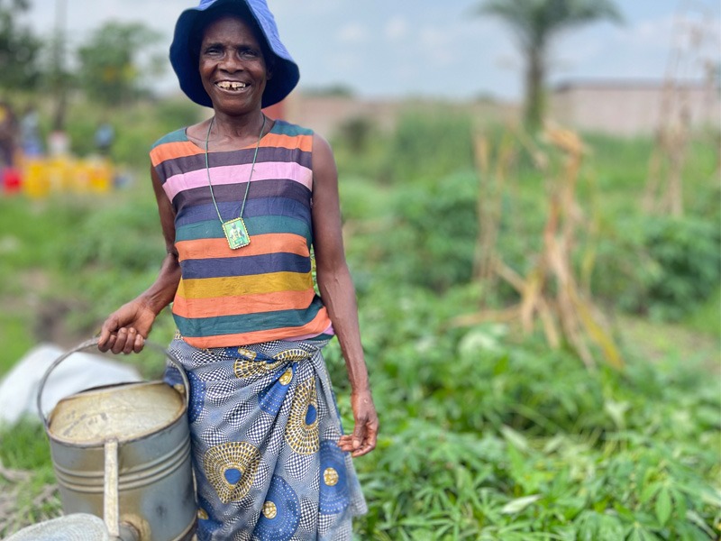 Eine afrikanische Frau steht mit einer Gießkanne vor einem Feld.