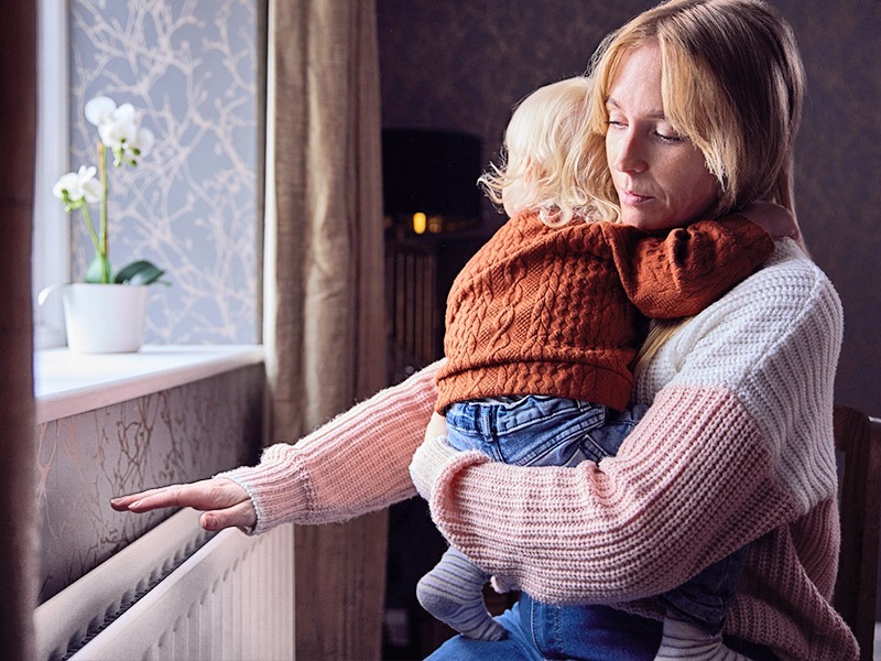 Eine Frau hält mit besorgtem Blick ihr kleines Kind im Arm und greift mit der anderen Hand über einen Heizkörper.