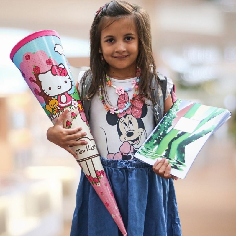 Ein Mädchen hält lächelnd eine Schultüte in der einen und ein Schulheft in der anderen Hand.