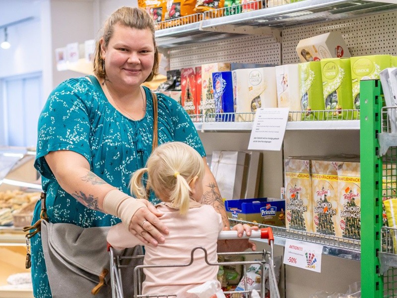 Eine Frau steht mit ihrer Tochter, die im Einkaufswagen sitzt, in einem Lebensmittelgeschäft und lächelt.