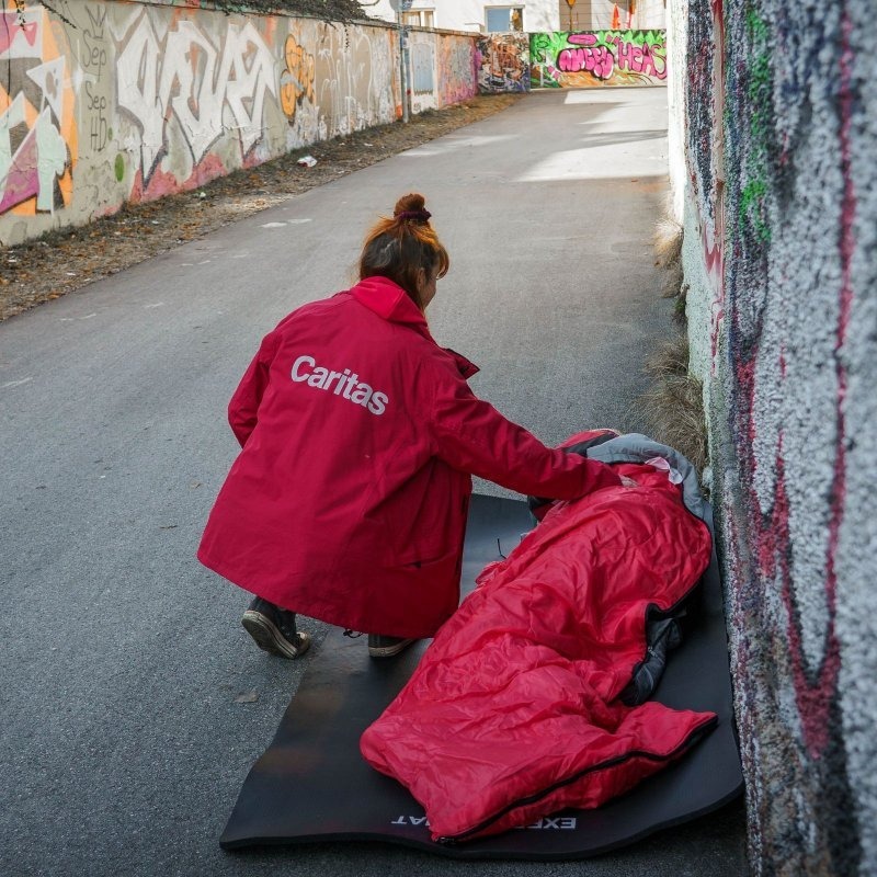 Eine Frau mit Caritas-Jacke kniet neben einem Menschen, der eingehüllt in einem Schlafsack im Freien liegt.
